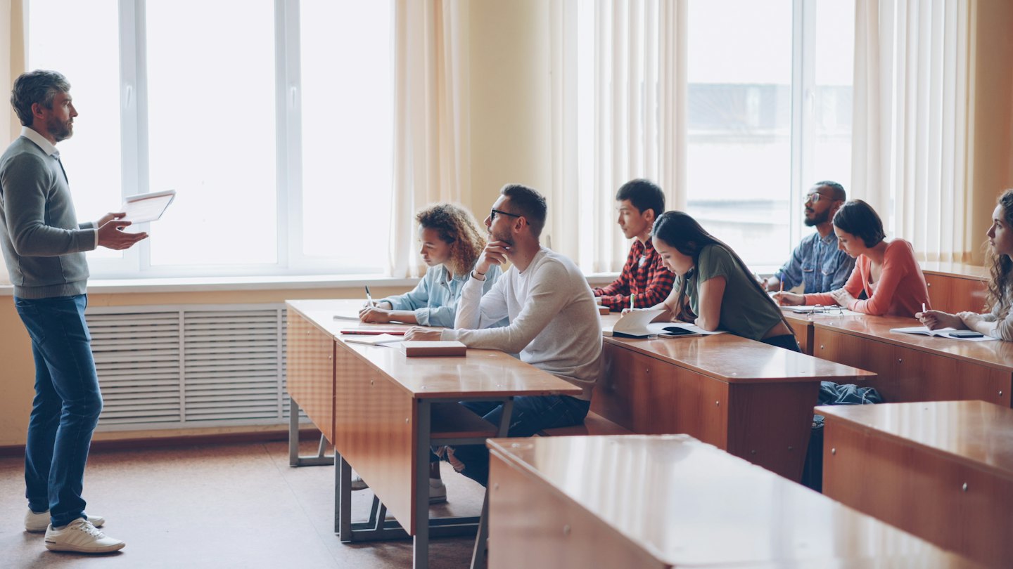 Teacher with tablet guides diverse students in a bright classroom.